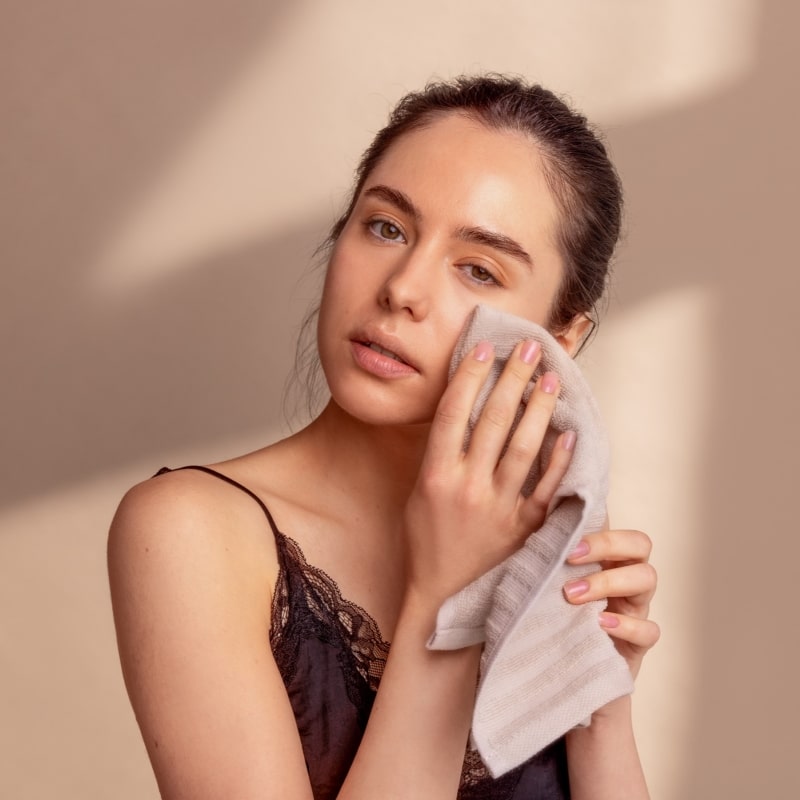 Woman cleaning her face with a towel against a beige background